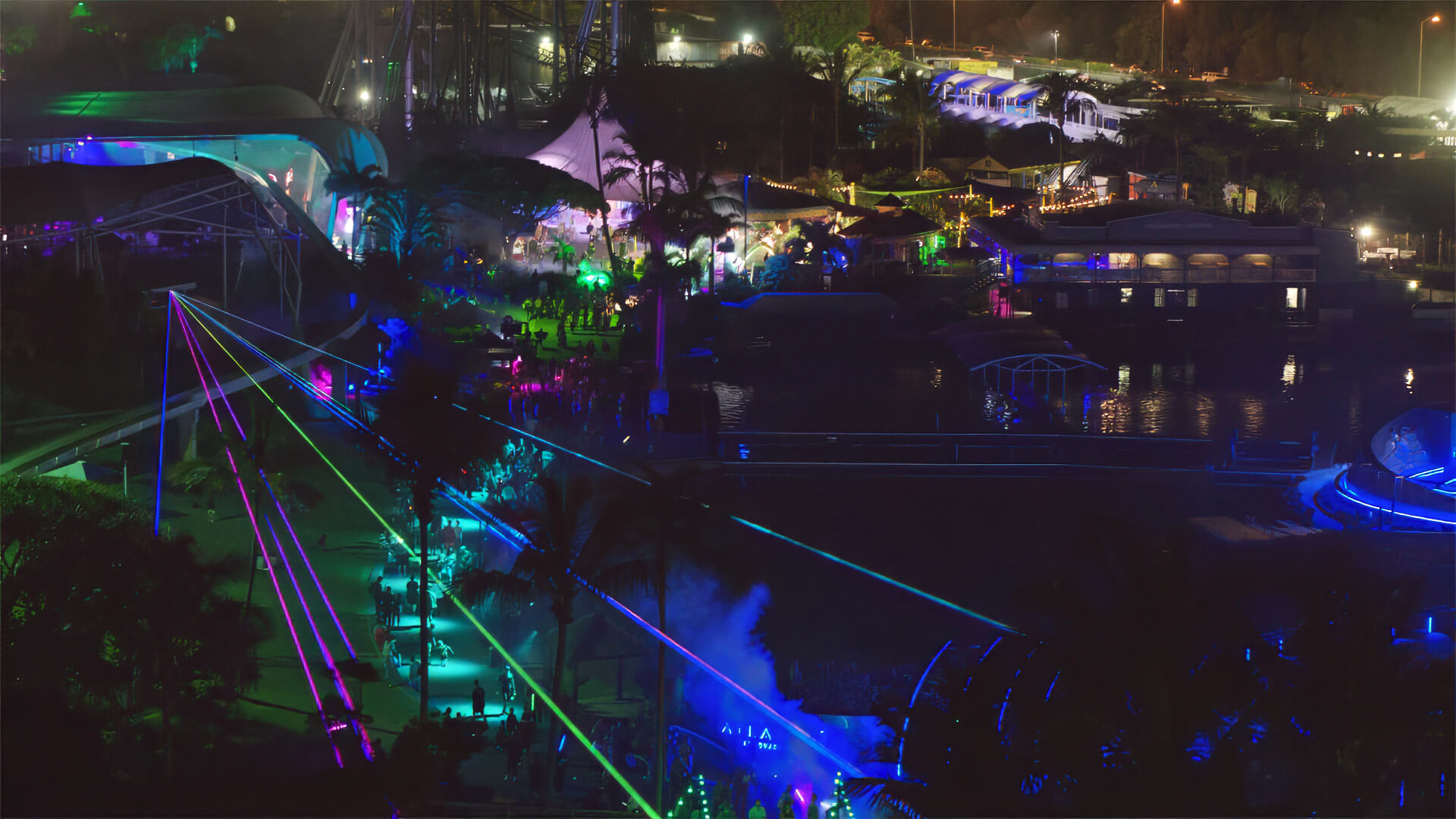 A nighttime scene at a park or festival is illuminated with colorful neon lights and laser beams, highlighting pathways, trees, and attractions, creating a lively and vibrant atmosphere.
