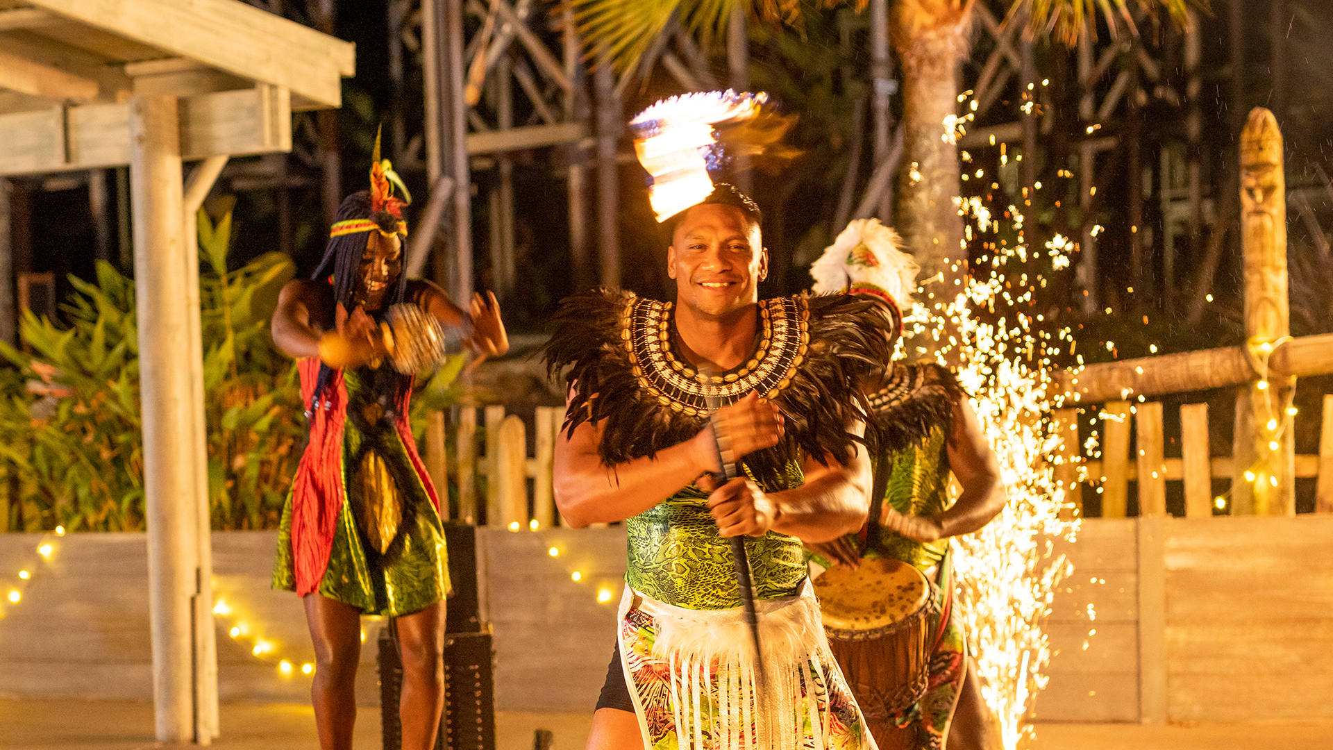 Three performers in colorful traditional attire dance at night; one smiles while spinning a staff with fire on top, with sparks in the background and tropical plants surrounding the scene.