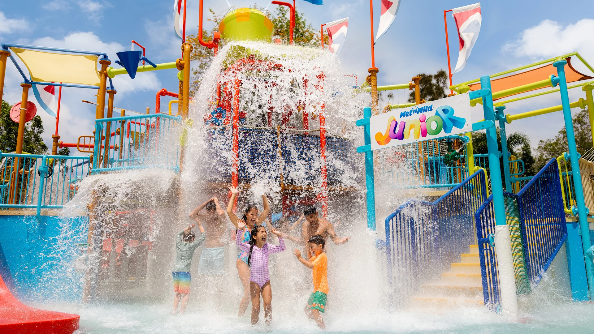 Children play under a large splash of water at a colorful outdoor water park, surrounded by slides, stairs, and flags. A sign reads “Junior” near the play structure. The scene is lively and fun.