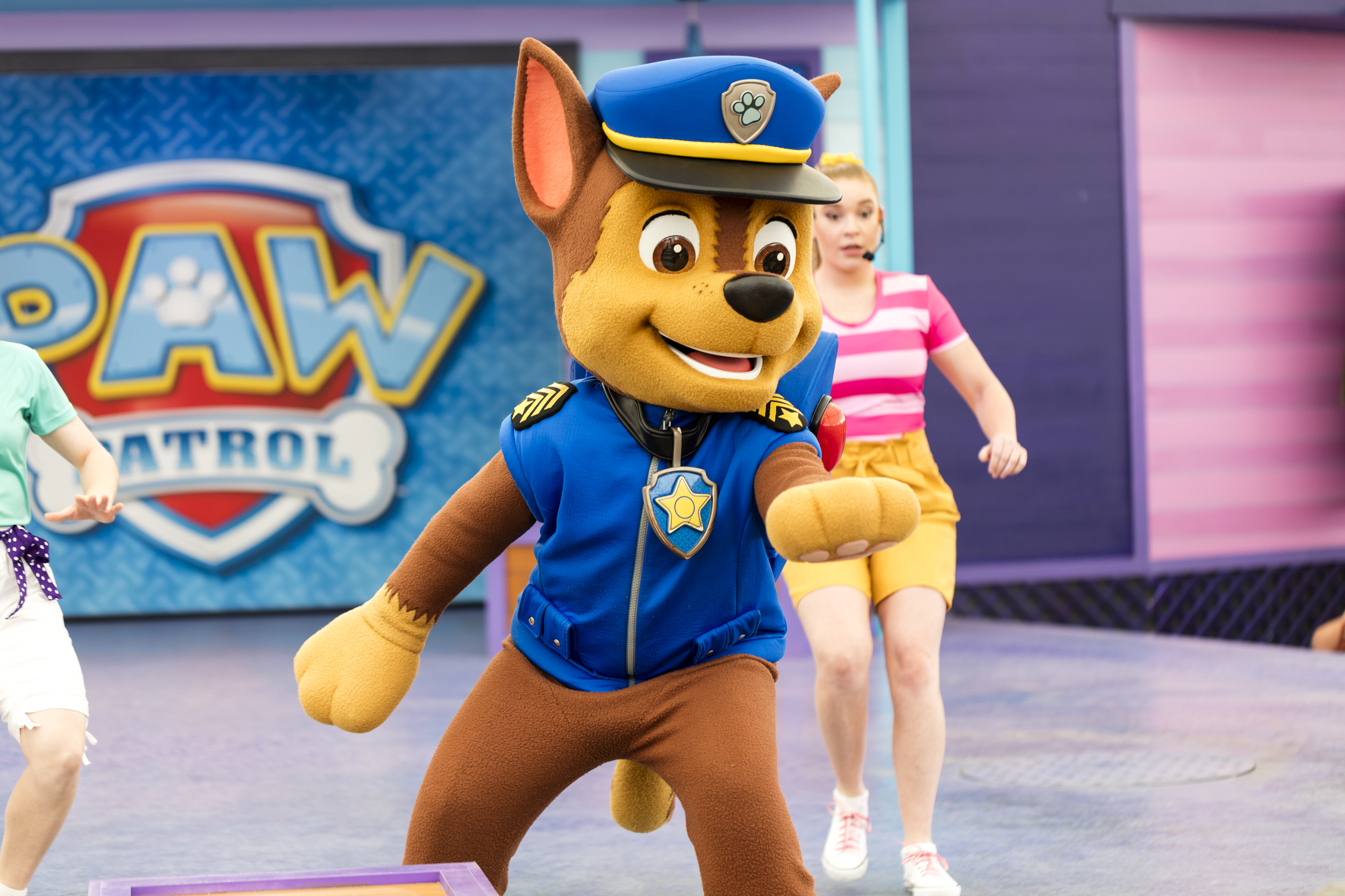 A performer in a Chase costume from PAW Patrol dances on stage with people in the background.