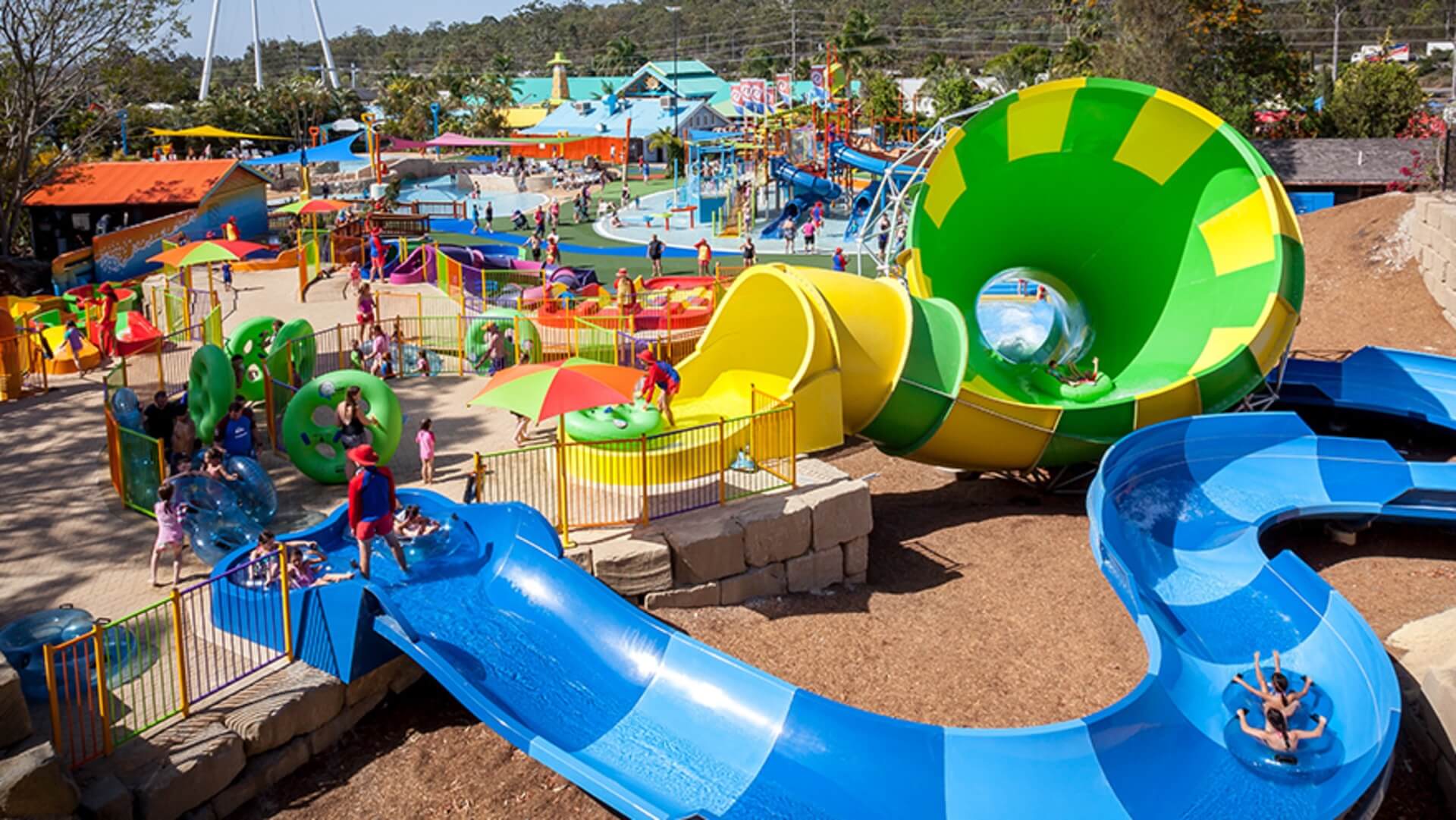 A colorful outdoor water park with winding slides, splash pads, and pools. Children and adults are enjoying various water attractions under sunny skies, surrounded by bright structures and greenery.