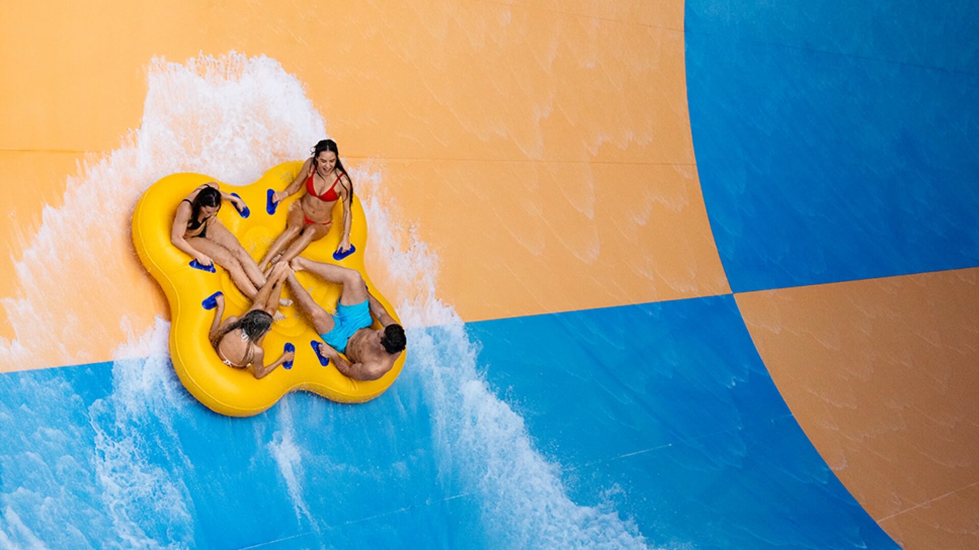 Group of friends riding a yellow raft down the Tornado water slide at Wet’n’Wild, surrounded by splashing water on bright orange and blue slide walls