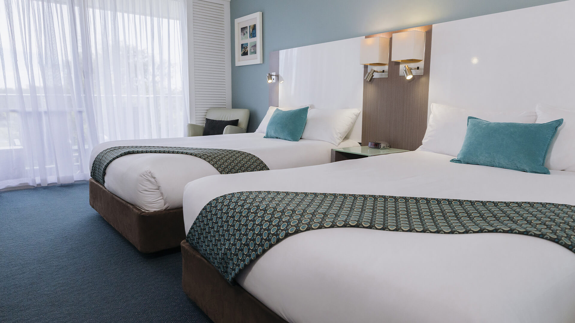 A hotel room with two neatly made double beds, blue accent pillows, and throws. A chair sits by a window with sheer curtains, and framed art decorates the wall.