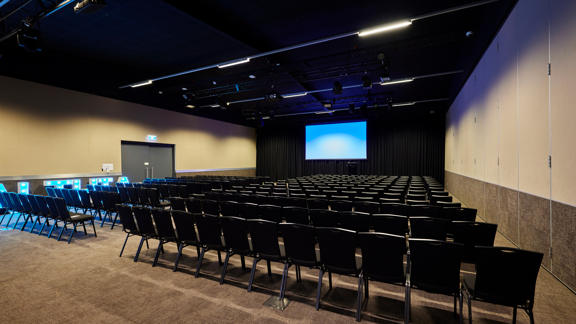 Rows of empty black chairs face a large projector screen in a modern, well-lit conference room with beige walls and dark ceiling.