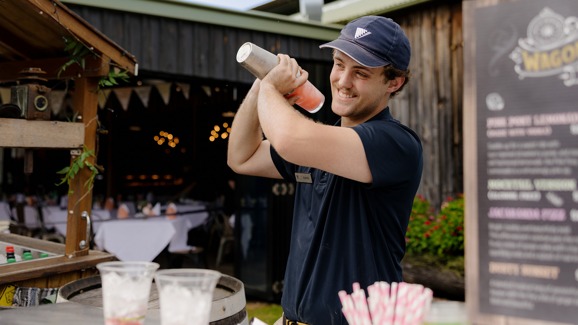 A smiling bartender wearing a navy shirt and cap shakes a cocktail shaker behind an outdoor bar, with drinks, straws, and a menu board visible in the foreground.