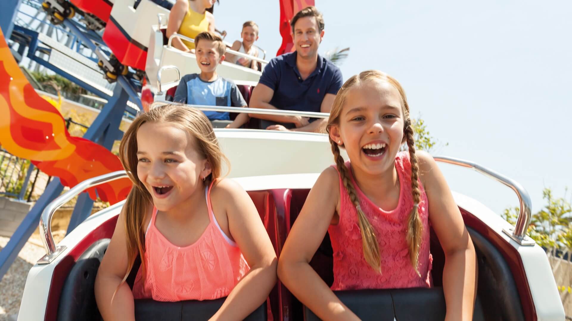 Two young girls and several people behind them ride a roller coaster outdoors on a sunny day, smiling and holding onto safety bars.