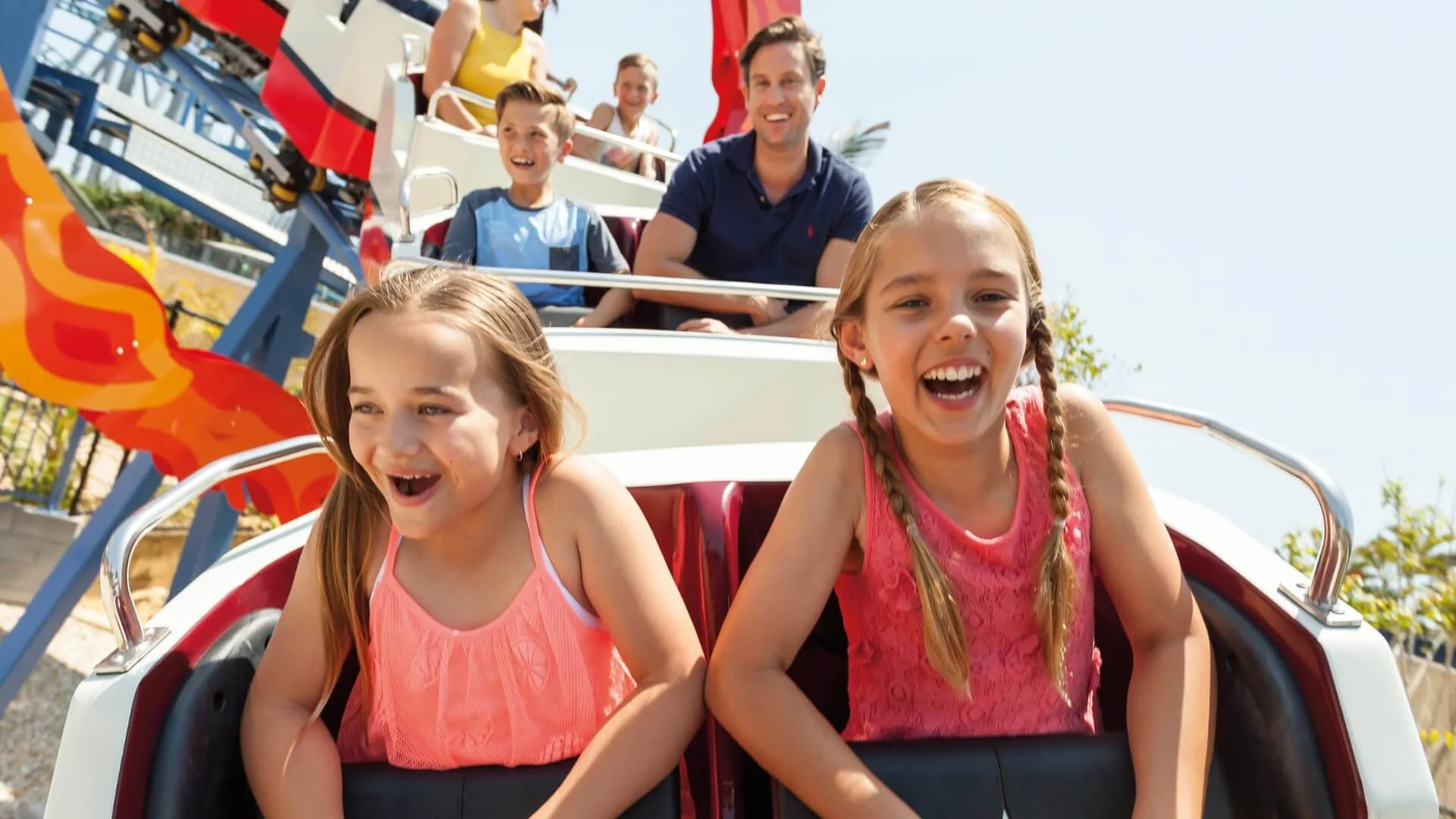 Two young girls and several people behind them ride a roller coaster outdoors on a sunny day, smiling and holding onto safety bars.
