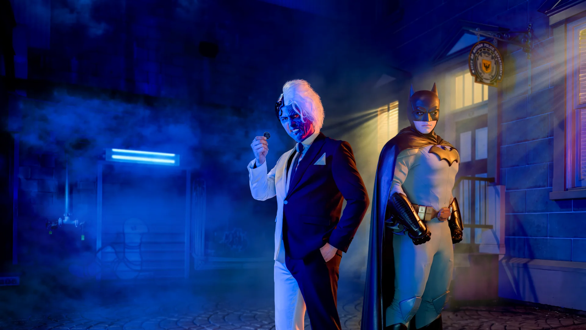Two costumed individuals stand in a dimly-lit alley: one in a white suit with a blue face holding a mask, and the other dressed as Batman, surrounded by blue and orange light.