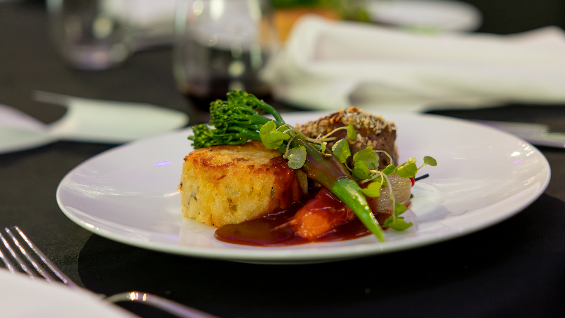 A plated gourmet meal featuring a round potato cake, a piece of meat with sauce, broccolini, carrots, and microgreens, set on a white plate with a fork and wineglass in the background.