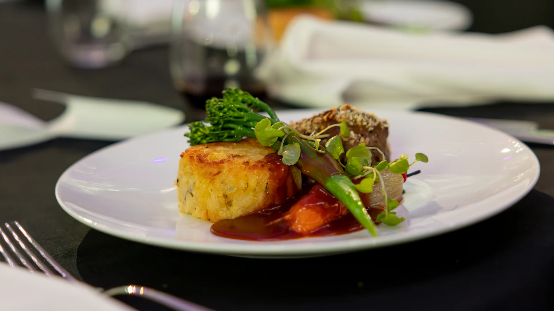 A plated gourmet meal featuring a round potato cake, a piece of meat with sauce, broccolini, carrots, and microgreens, set on a white plate with a fork and wineglass in the background.