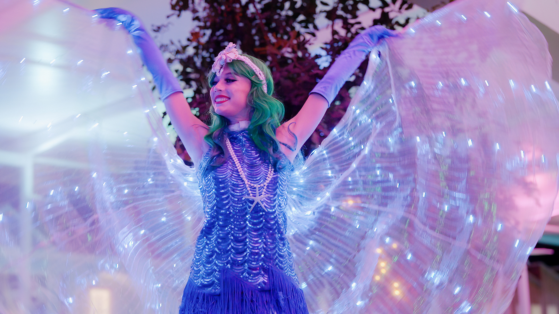 A performer in a blue fringed costume with green hair and glowing wings poses with arms raised, illuminated by purple and blue lights.