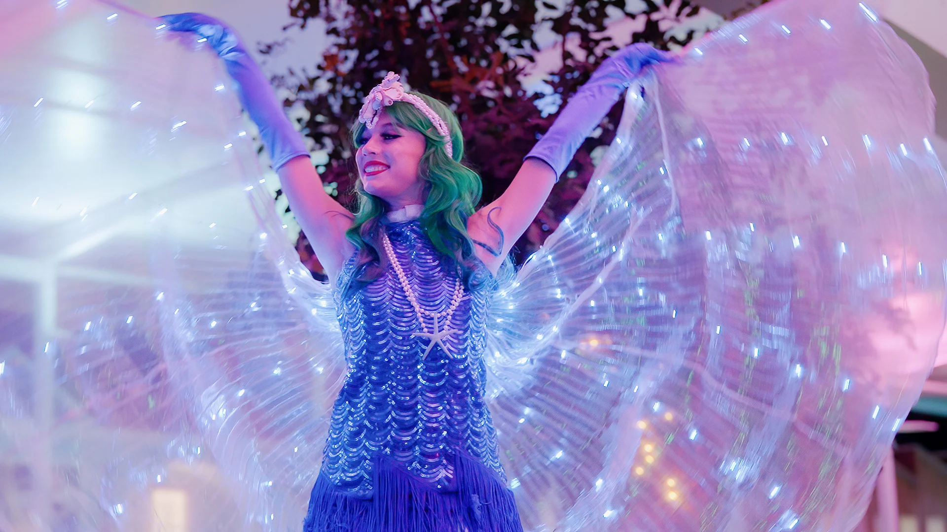 A performer in a blue fringed costume with green hair and glowing wings poses with arms raised, illuminated by purple and blue lights.