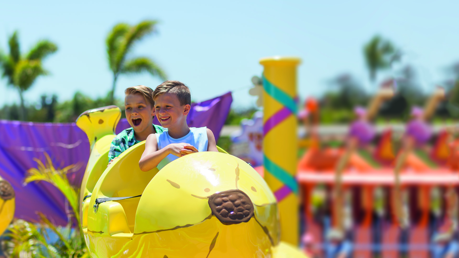 Two boys ride a yellow amusement park ride shaped like an animal, with palm trees and colorful decorations in the background.
