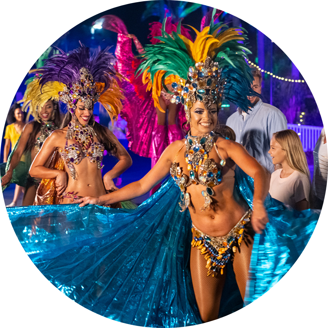 Two women in colorful feathered costumes and elaborate headdresses dance at a lively outdoor event, holding blue fabric wings, while onlookers, including a young girl, watch and smile.