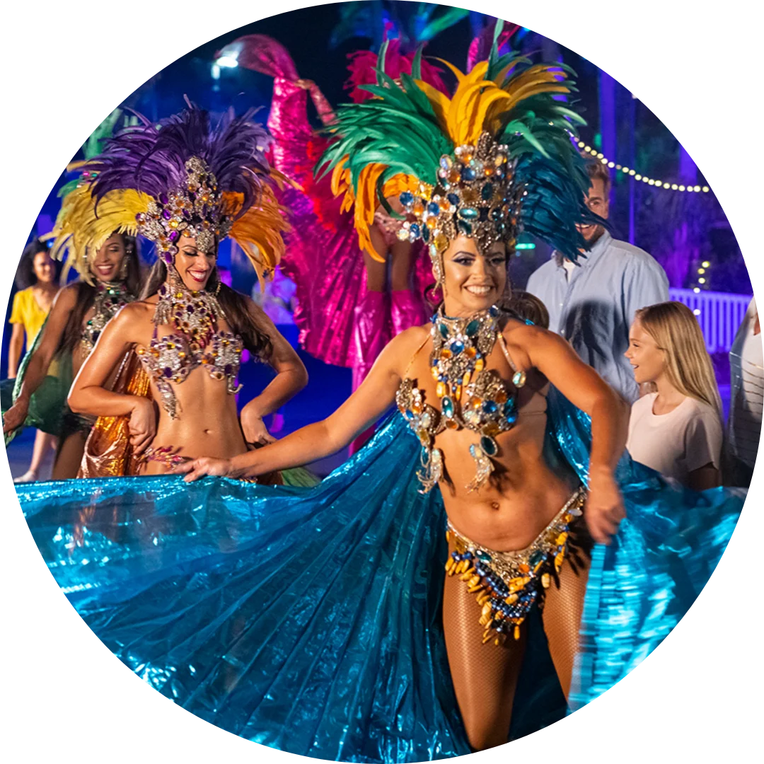 Two women in colorful feathered costumes and elaborate headdresses dance at a lively outdoor event, holding blue fabric wings, while onlookers, including a young girl, watch and smile.