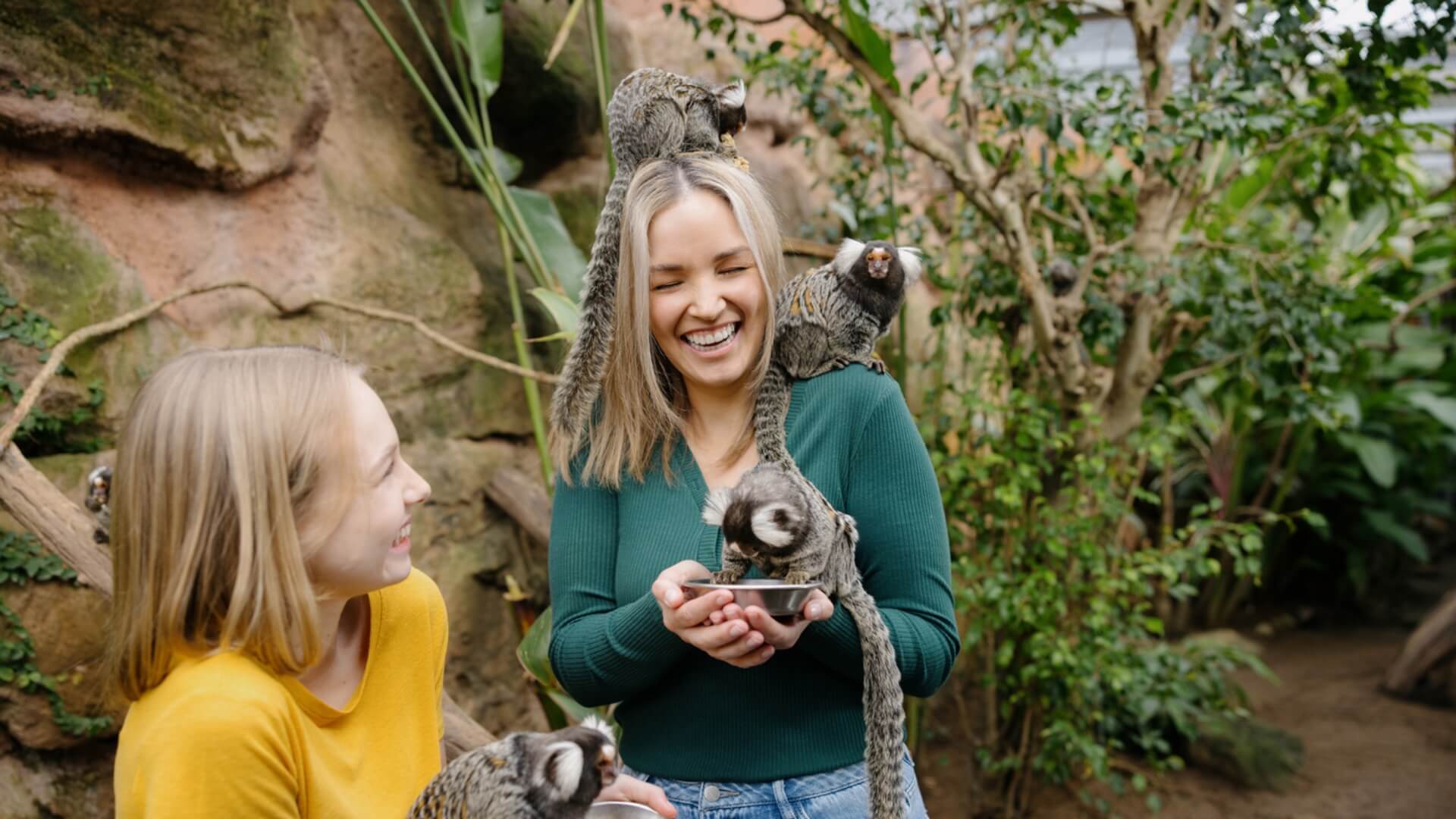 Two women stand in a lush, indoor enclosure, smiling as several small monkeys climb on and around them.