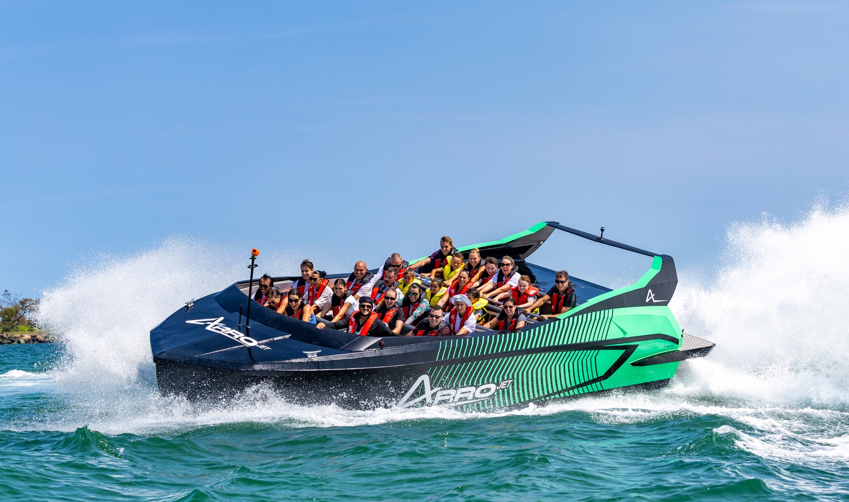 A group of people wearing life jackets ride a black and green speedboat on bright blue water, creating large splashes under a clear blue sky.