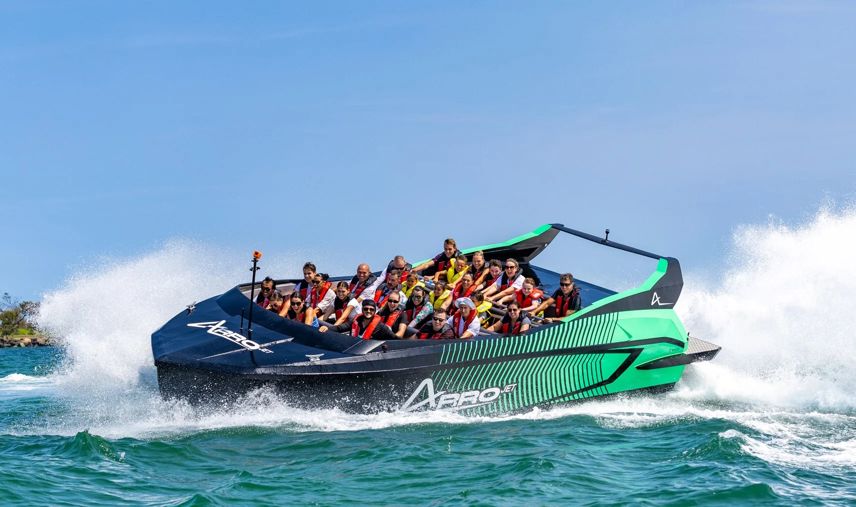 A group of people wearing life jackets ride a black and green speedboat on bright blue water, creating large splashes under a clear blue sky.