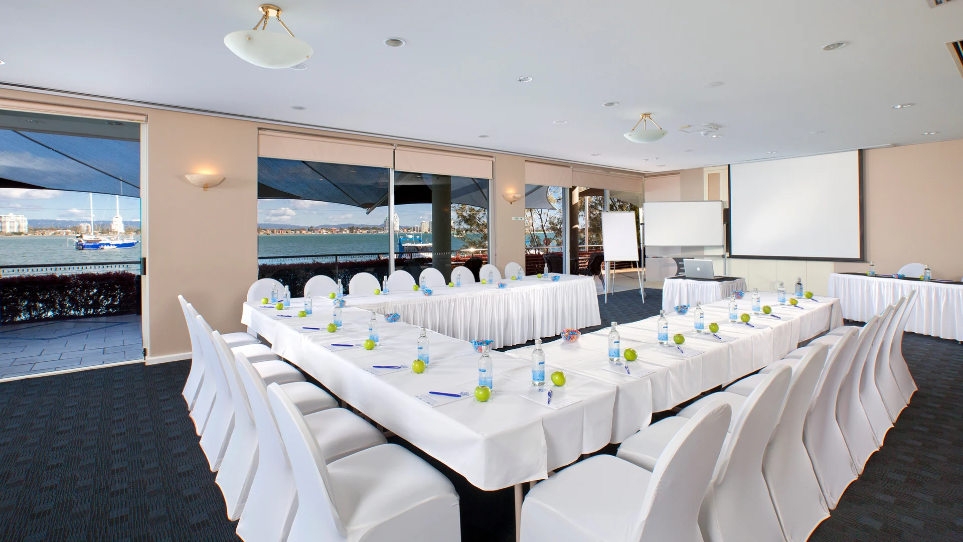 A bright conference room with large windows overlooking water, set up with U-shaped tables, white chairs, notepads, pens, and green water bottles, ready for a meeting or presentation.