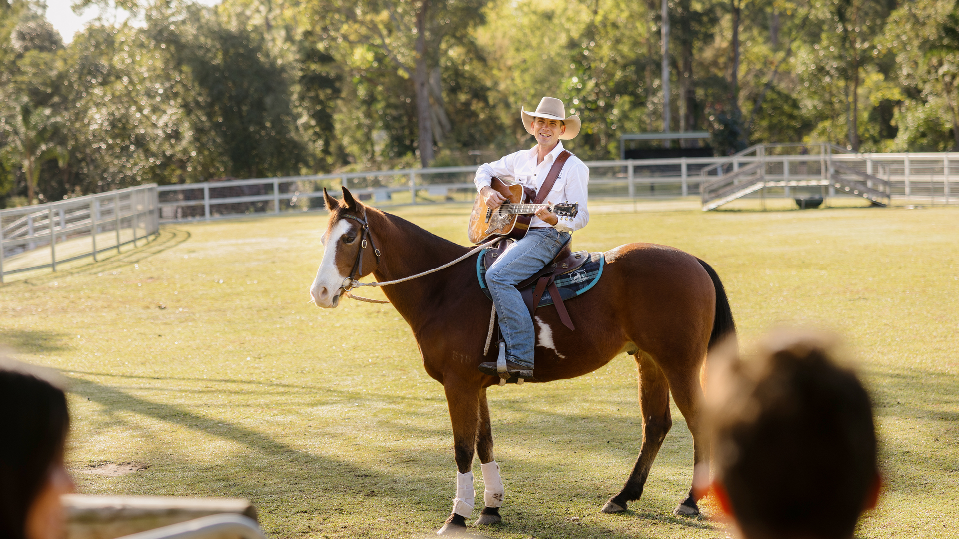 A person wearing a cowboy hat plays an acoustic guitar while sitting on a brown horse in a sunny, fenced pasture with trees in the background. Two people watch in the foreground.