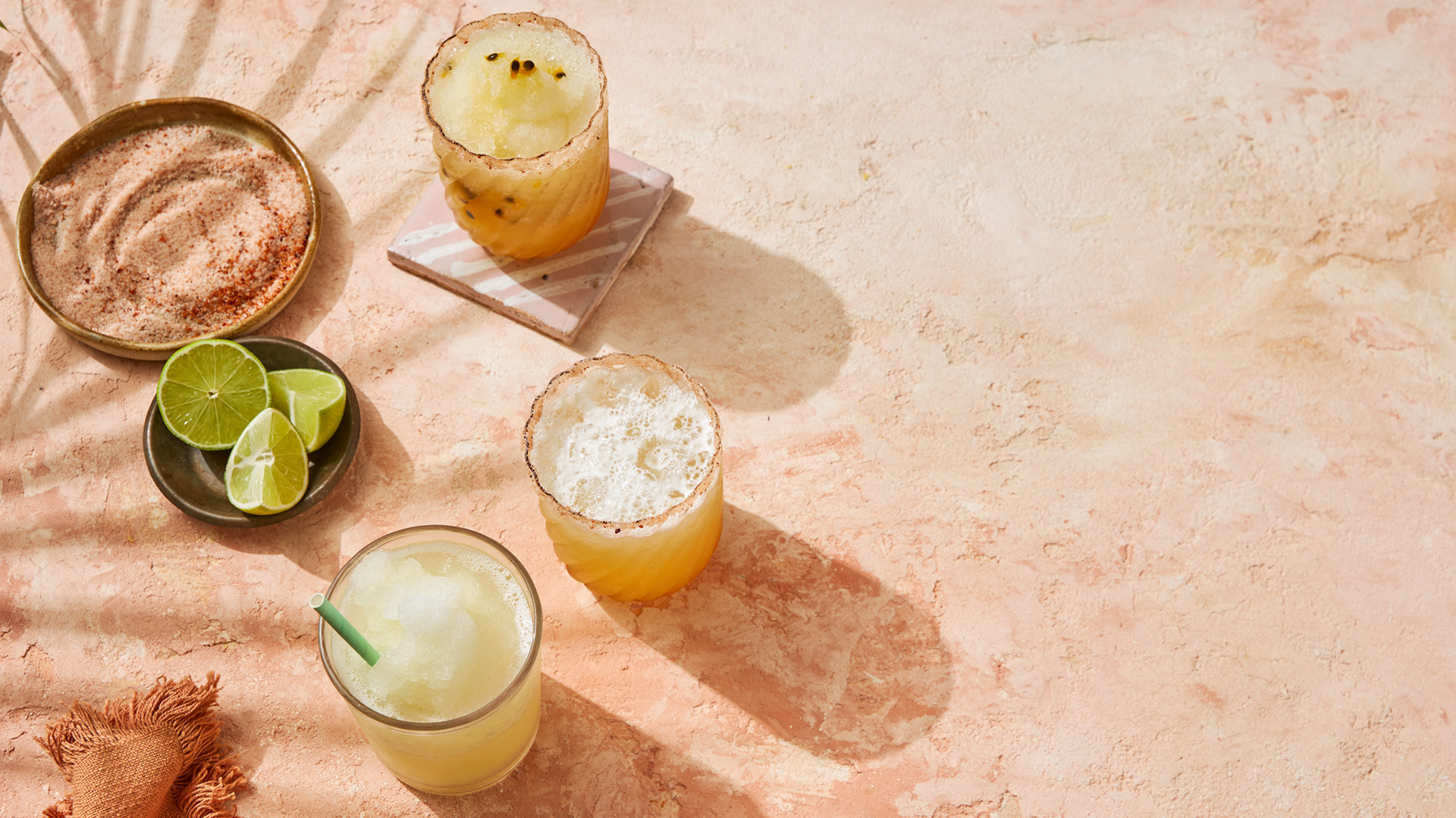 Three glasses of margaritas, a bowl of sliced limes, and a plate of chili salt are arranged on a sunlit peach-colored surface with a leafy shadow cast across the scene.