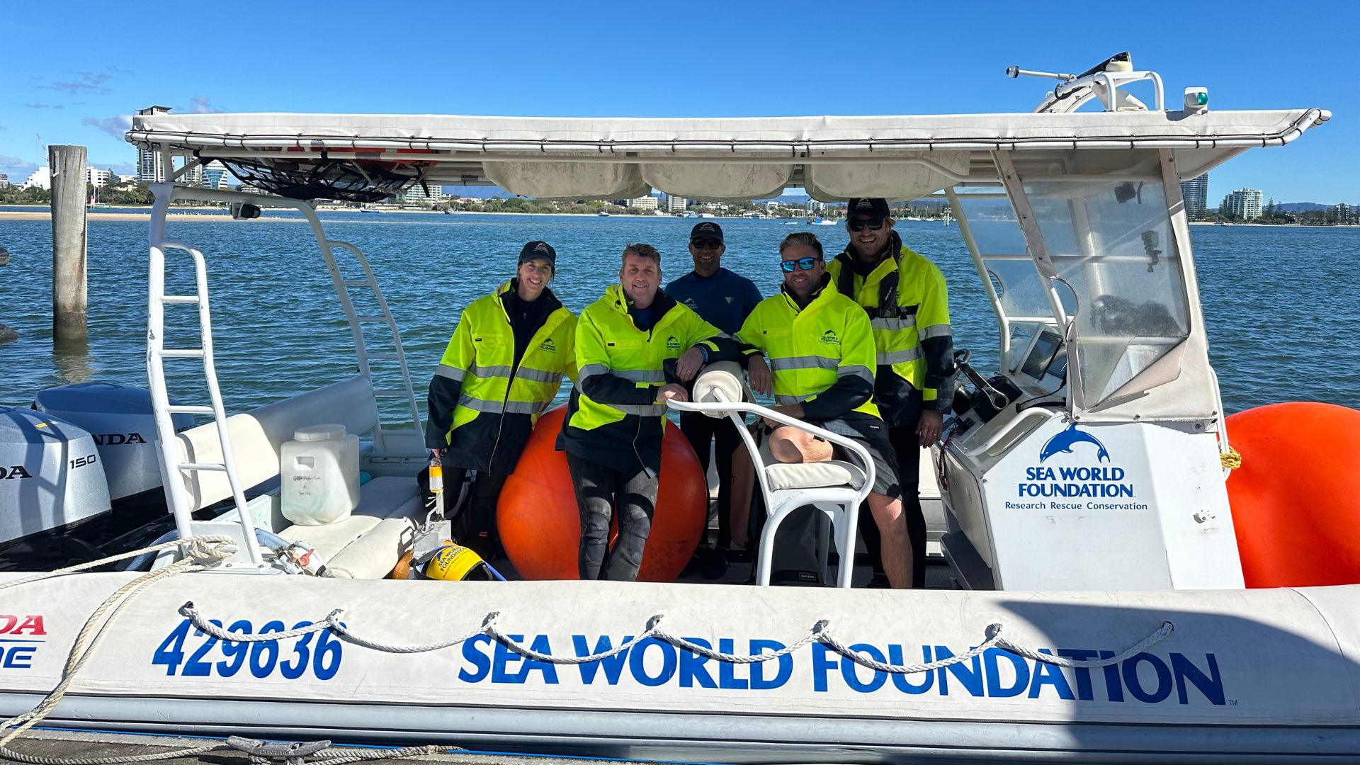 Six people wearing yellow safety jackets sit and stand on a Sea World Foundation rescue boat docked in calm water, with buildings and blue sky visible in the background.