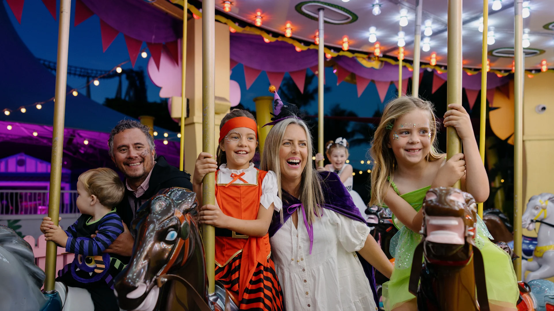 A family with young children, some in costumes, smiles and enjoys riding a brightly lit carousel at night.