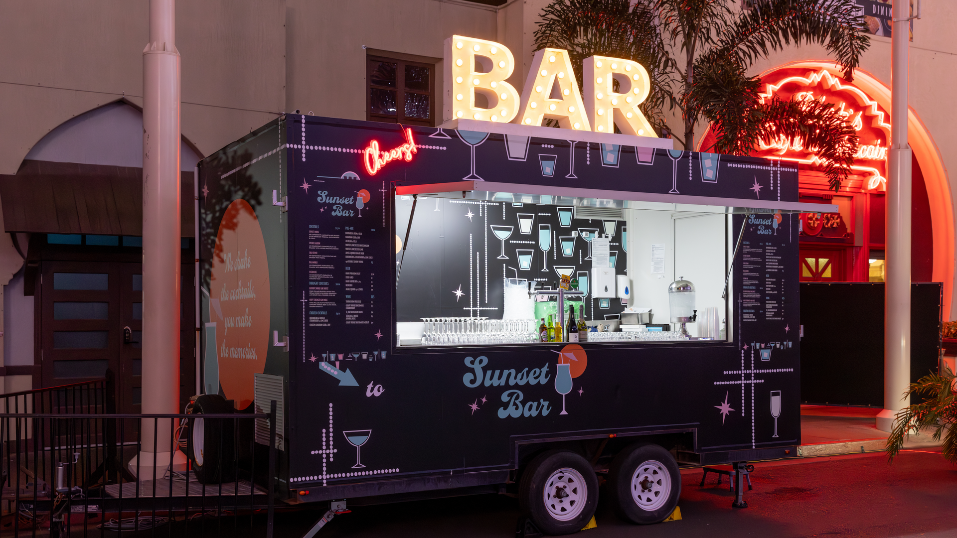 A black mobile bar trailer labeled "Sunset Bar" is parked outdoors at night, illuminated by a large, glowing "BAR" sign on top. Neon signs and palm trees are visible in the background.