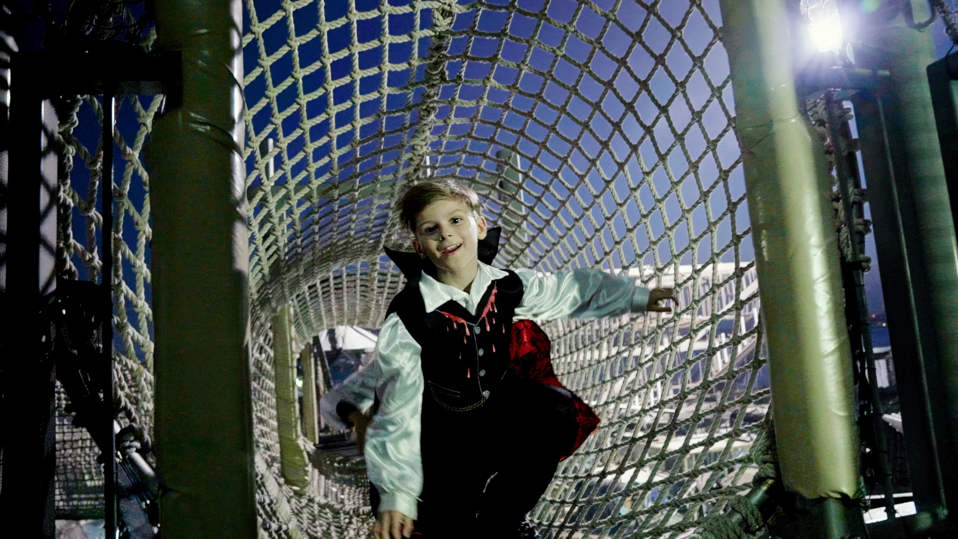 A child in a costume crawls through a rope tunnel on a playground at night, illuminated by bright lights in the background.