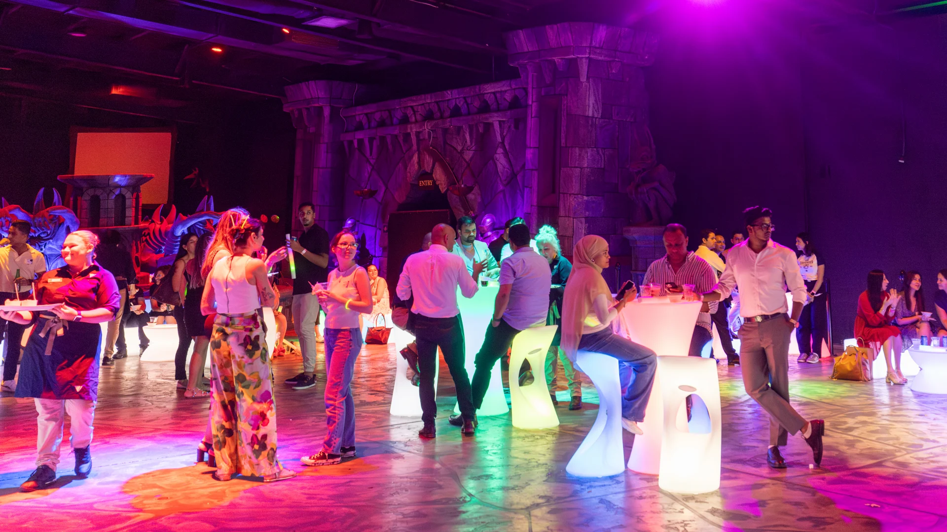 A group of people socialize and enjoy drinks in a colorful, dimly lit venue with glowing tables and medieval-style decor. The atmosphere is lively and festive.