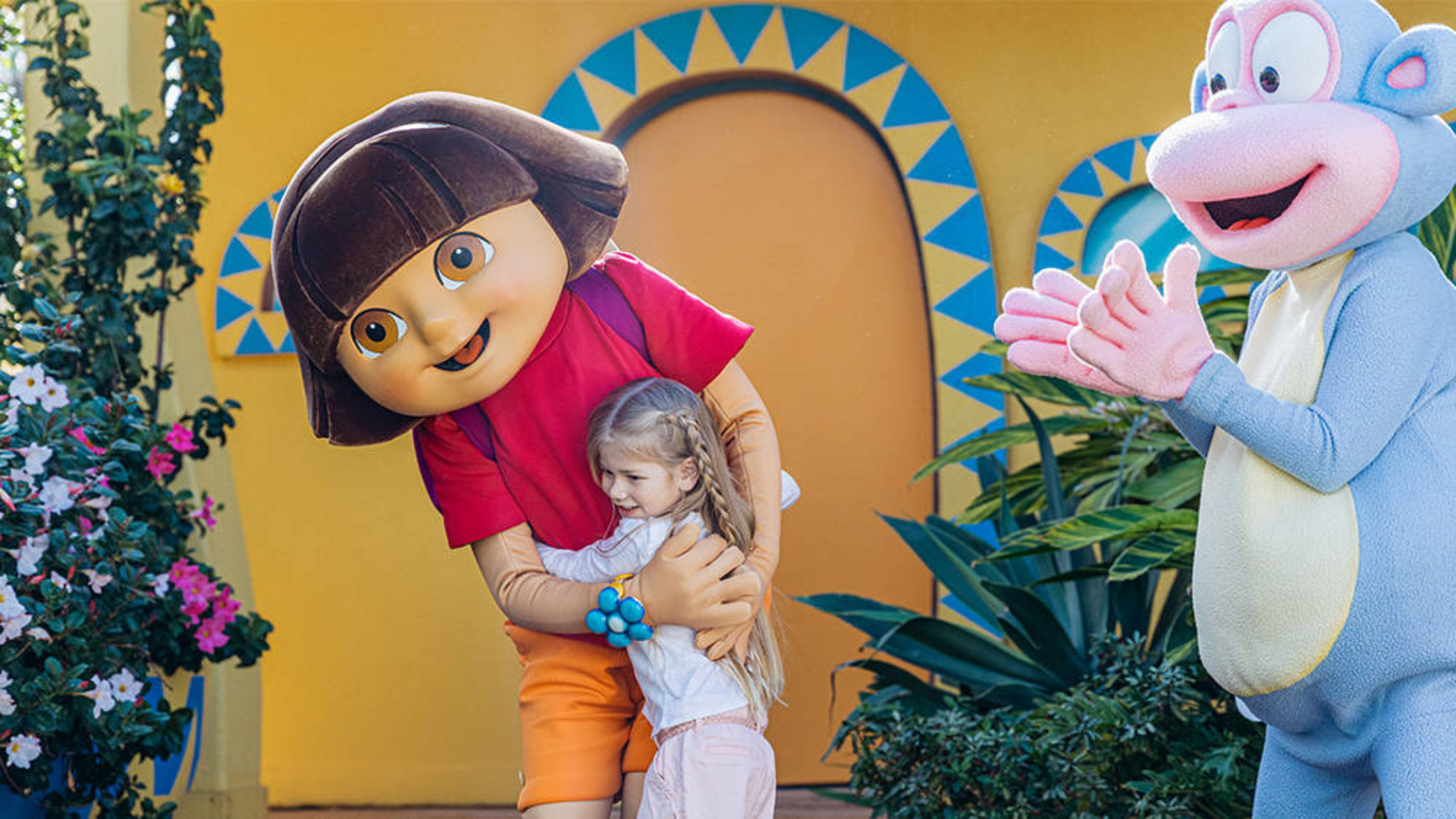 A child hugs a person in a Dora the Explorer costume while another person in a Boots the Monkey costume stands nearby, clapping.