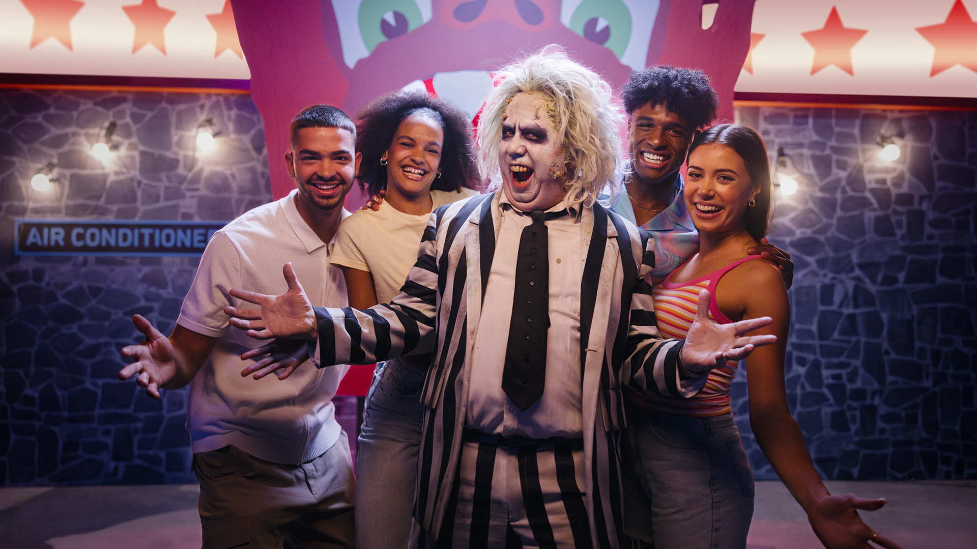 A person dressed as Beetlejuice in a black-and-white striped suit poses with four smiling young adults against a colorful, carnival-like background with star decorations and an "Air Conditioned" sign.