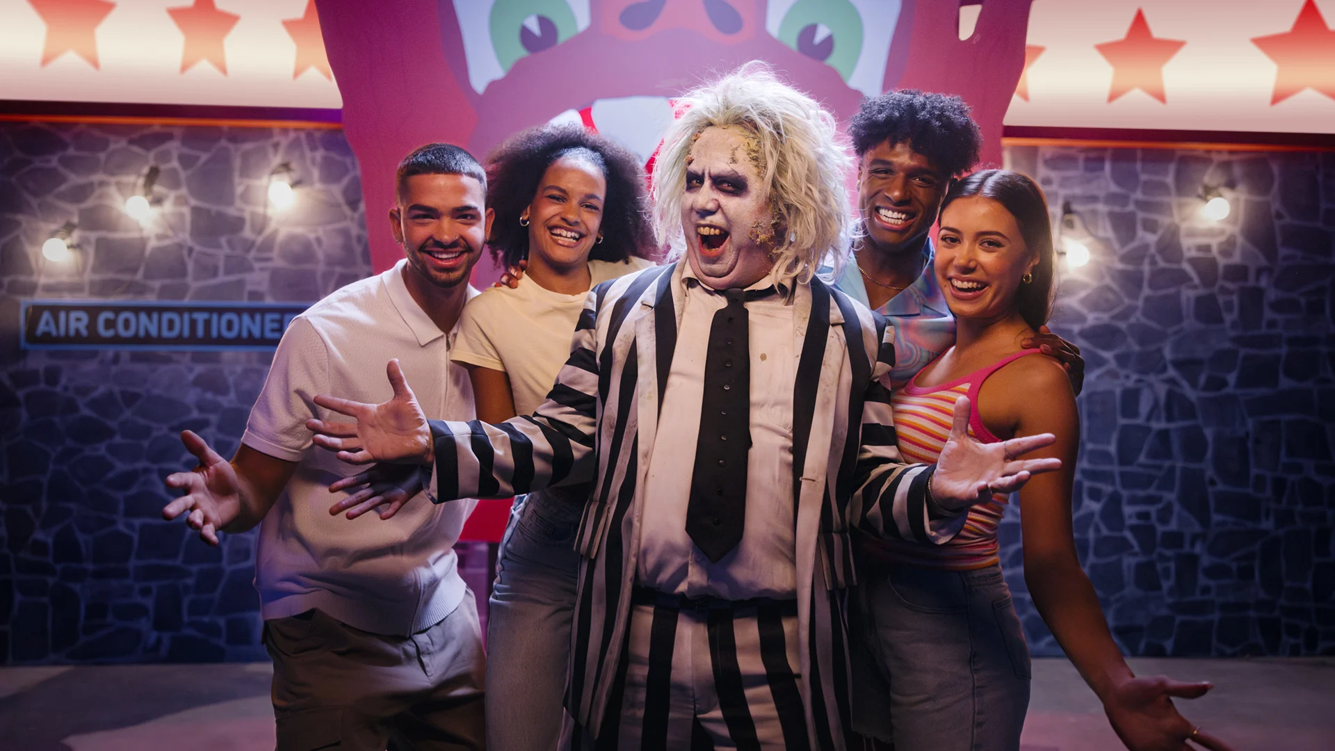 A person dressed as Beetlejuice in a black-and-white striped suit poses with four smiling young adults against a colorful, carnival-like background with star decorations and an "Air Conditioned" sign.