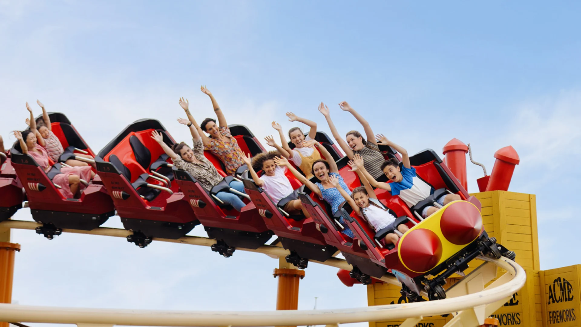 A group of people, including children and adults, ride a red roller coaster car with their hands up, smiling and enjoying the thrill under a clear blue sky.