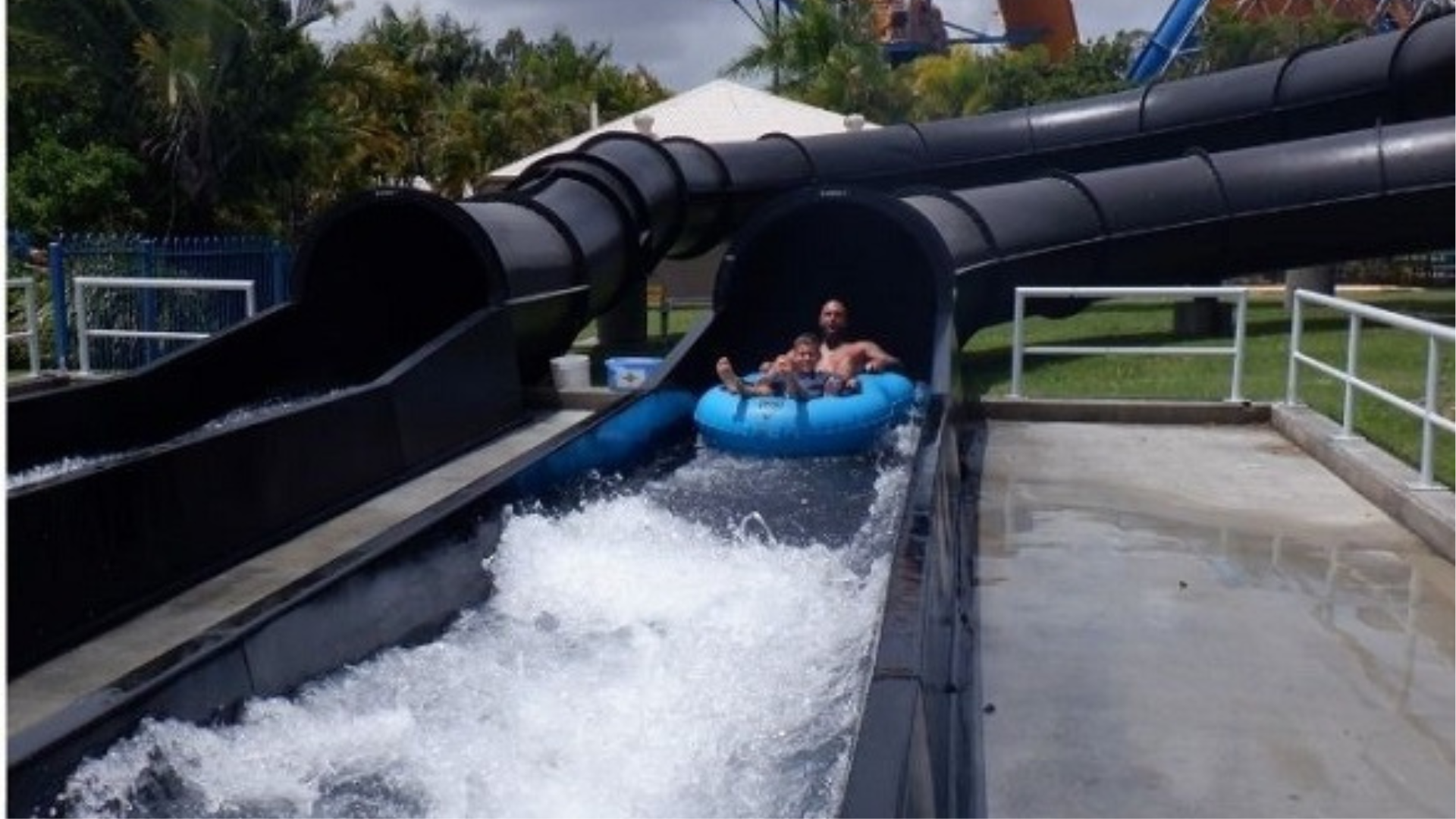 Two people ride a blue inflatable raft out of a black enclosed water slide, splashing into a pool at an outdoor water park surrounded by greenery and railings.