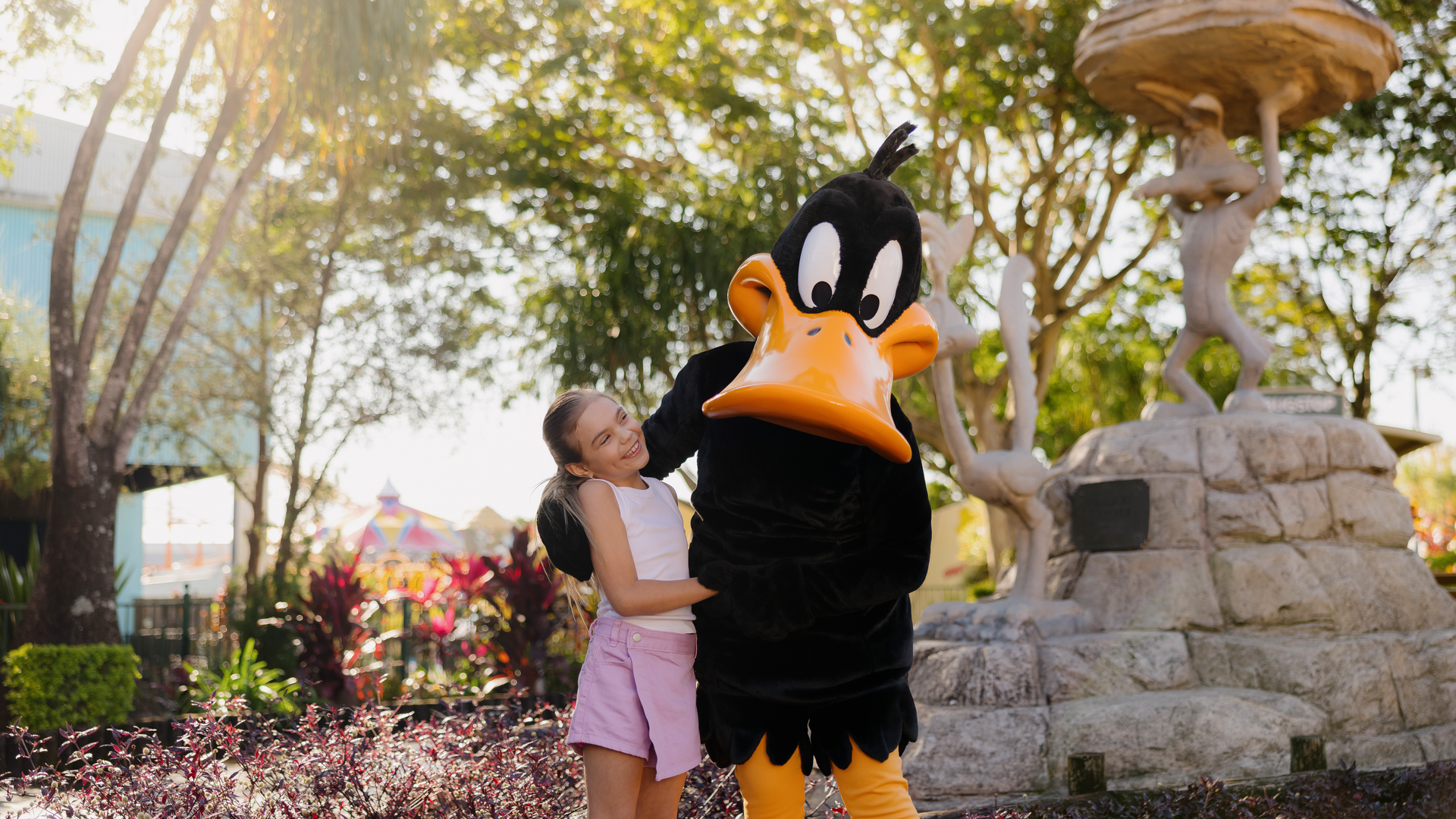 A smiling girl in light pink shorts and a t-shirt poses with a person in a Daffy Duck costume at an outdoor park, surrounded by trees, plants, and a stone fountain in the background.