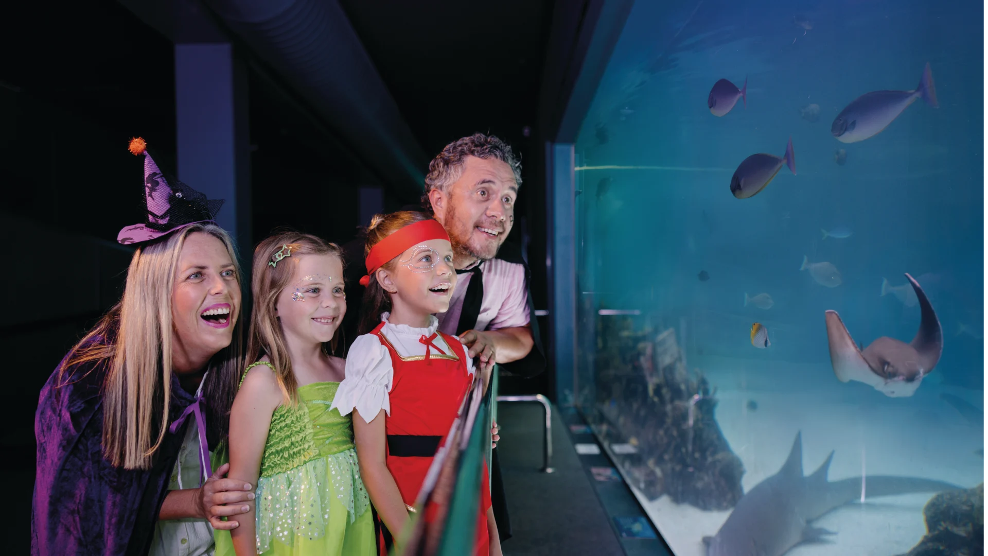 A family dressed in costumes smiles while looking at fish and sea creatures through the glass at an aquarium exhibit.