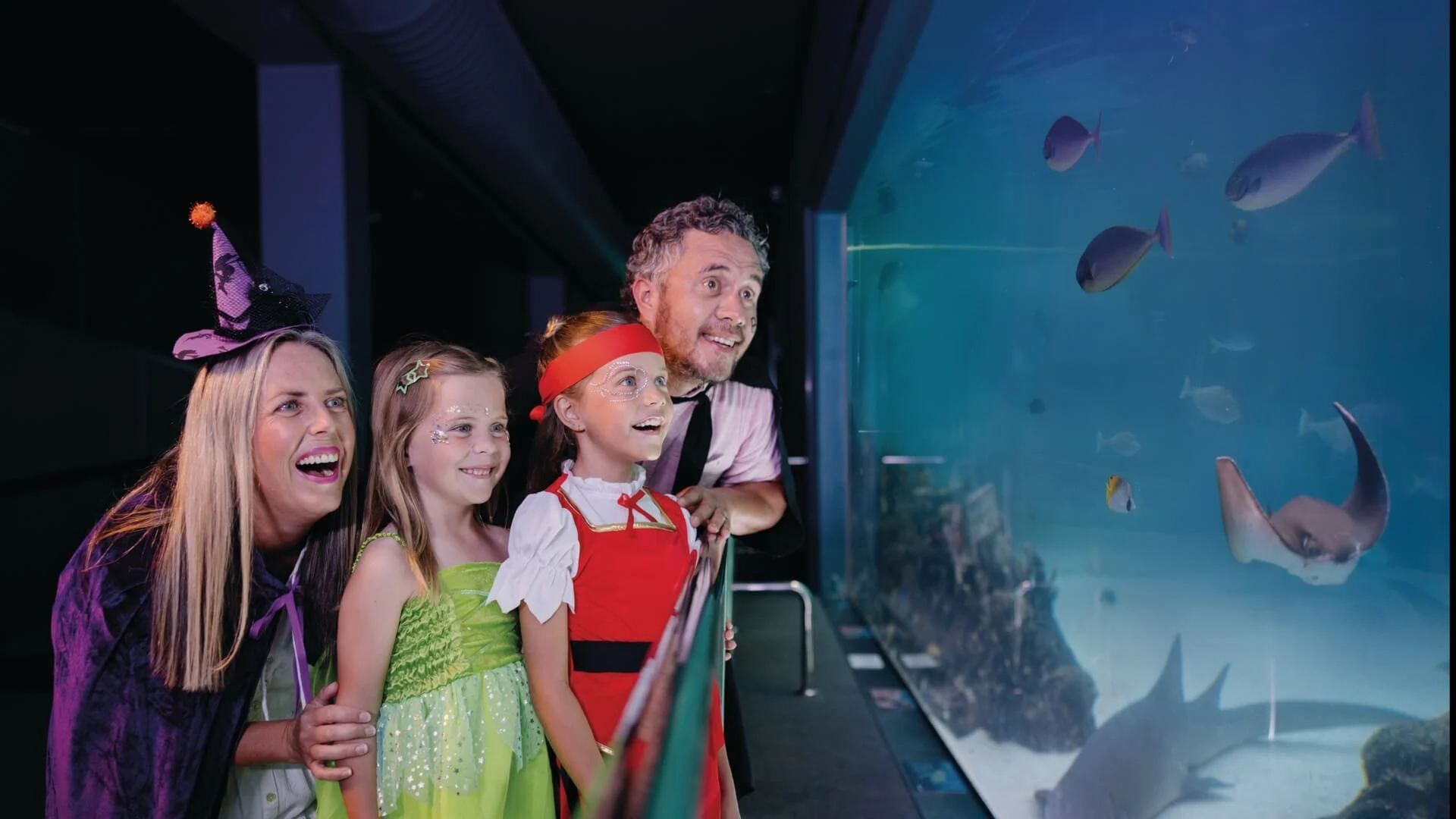 A family dressed in costumes smiles while looking at fish and sea creatures through the glass at an aquarium exhibit.