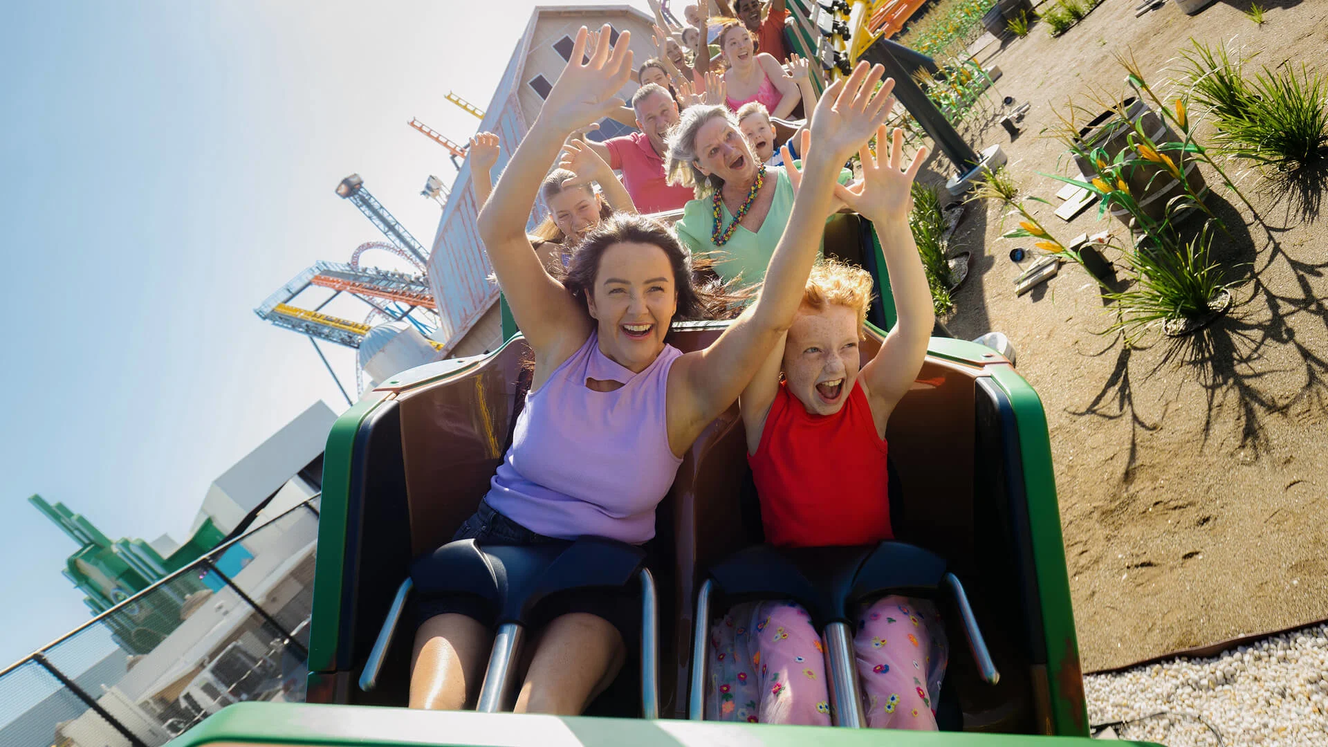 A woman and a child with wide smiles raise their arms as they ride a roller coaster, with other excited riders behind them and a sunny amusement park in the background.