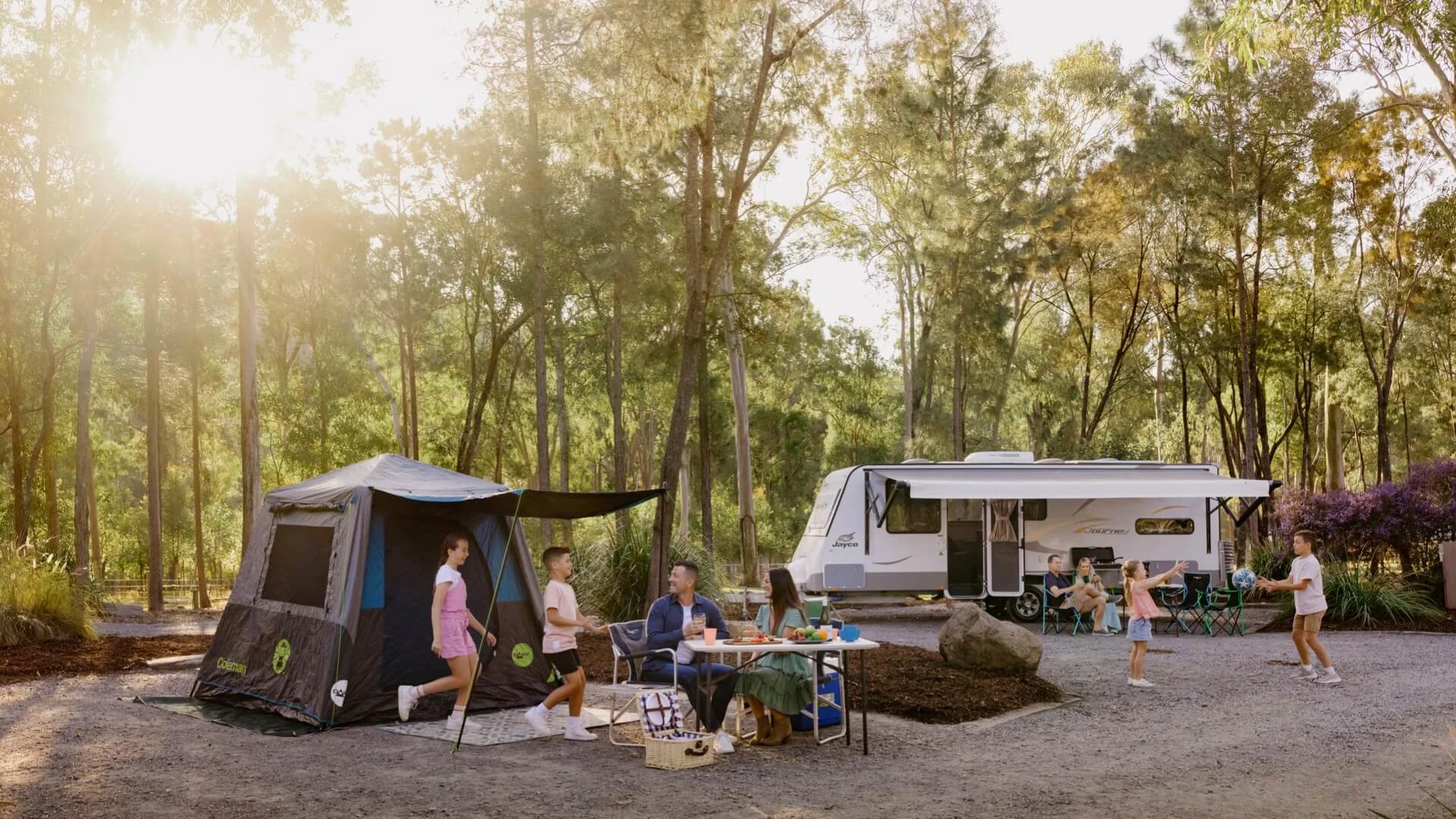 Families camping at Paradise Country with a caravan and tent set up among tall trees, children playing while adults relax at a picnic table in the sunlight