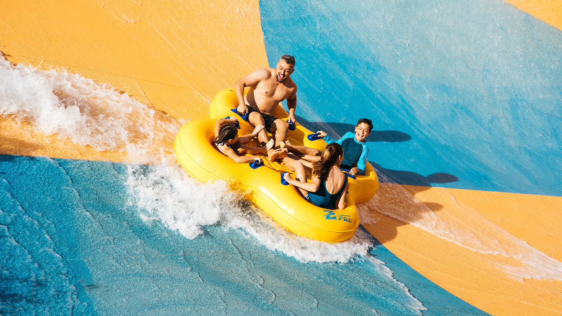 Four people ride together in a yellow inflatable raft down a colorful blue and orange water slide, surrounded by splashing water, enjoying a fun moment at a water park.