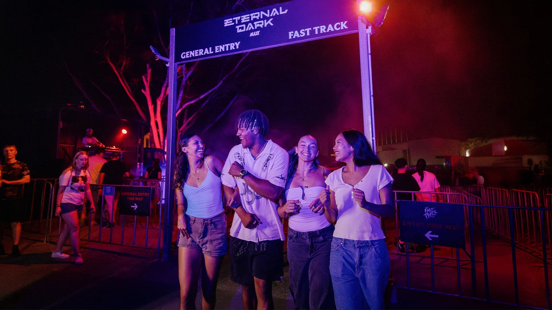 Four people smile and walk together under a sign reading “Eternal Dark” at night, surrounded by purple and red lights, with others and fencing in the background.
