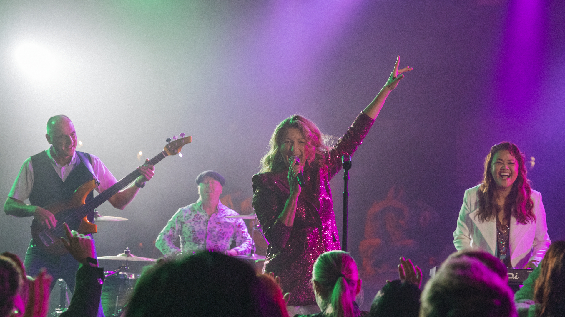 A band performs energetically on stage under colorful lights; the lead singer, in a sparkly dress, raises one arm while singing, with a bassist, drummer, and keyboardist playing beside her.