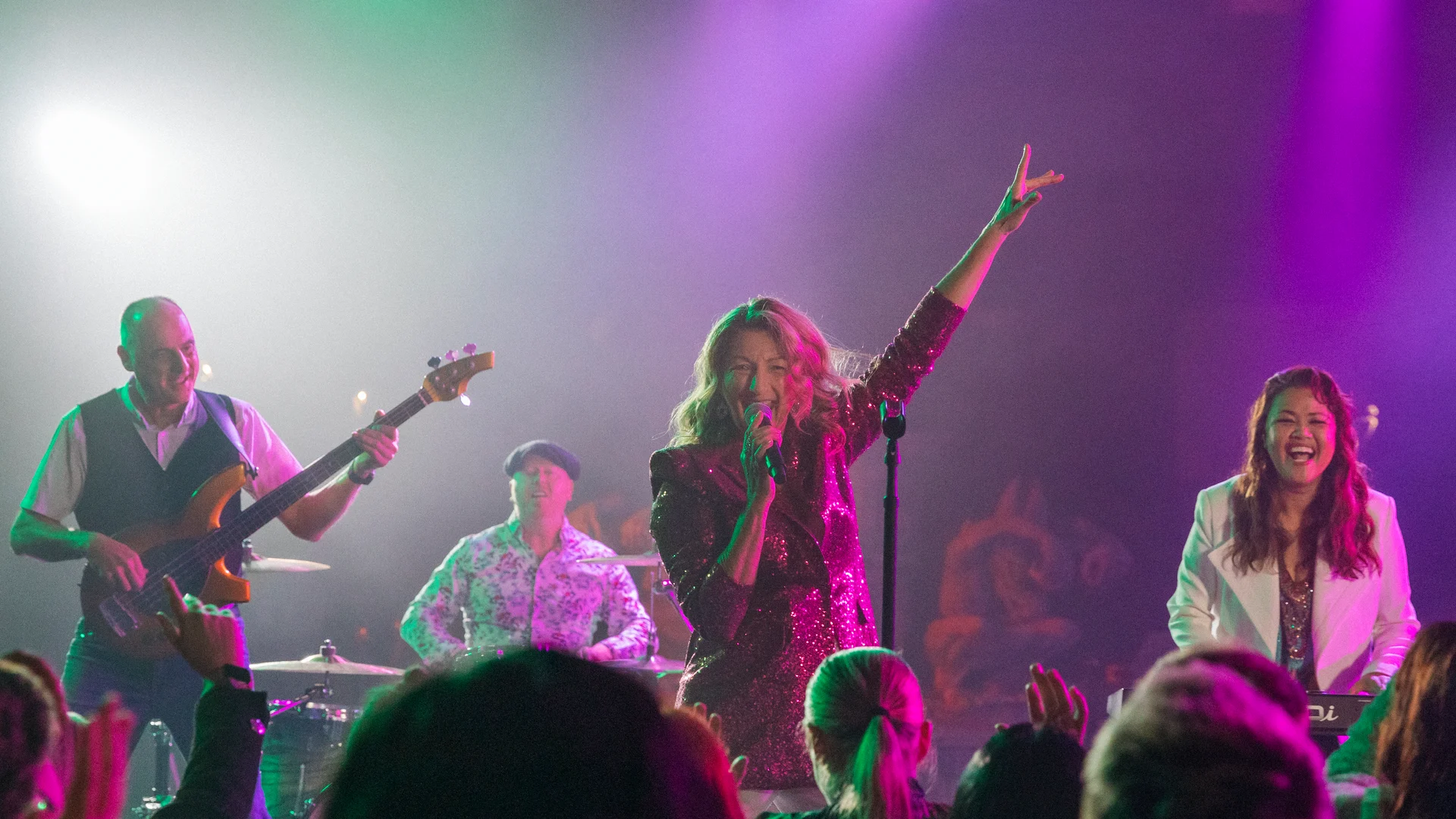 A band performs energetically on stage under colorful lights; the lead singer, in a sparkly dress, raises one arm while singing, with a bassist, drummer, and keyboardist playing beside her.