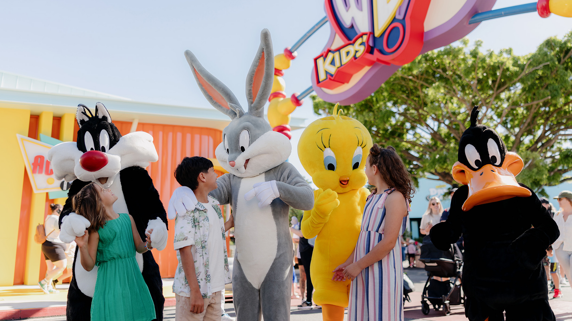 People stand outdoors with characters dressed as Sylvester, Bugs Bunny, Tweety, and Daffy Duck from Looney Tunes at a colorful theme park entrance under a sunny sky.