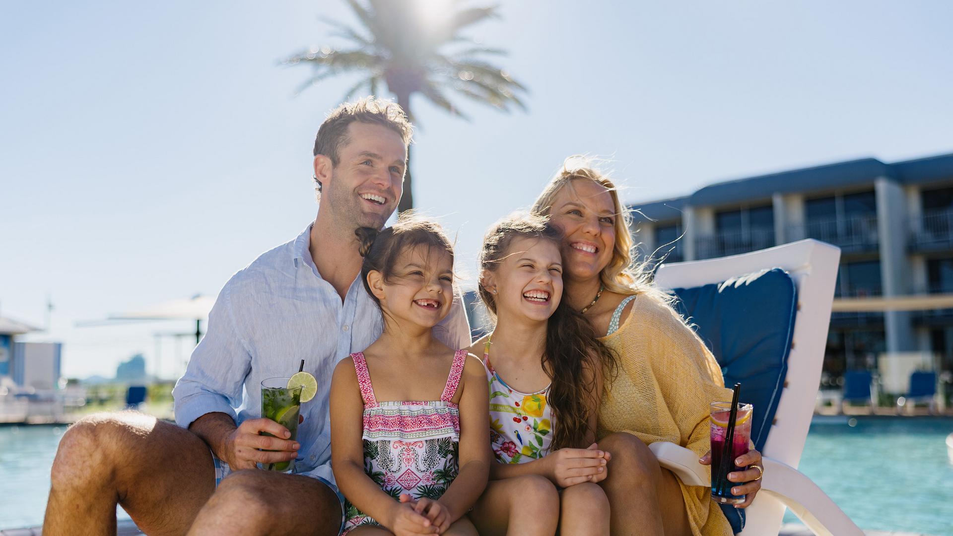 A smiling family of four sits on poolside lounge chairs, enjoying drinks on a sunny day. The parents and two young daughters are dressed in summer clothes, with a palm tree and a resort building in the background.