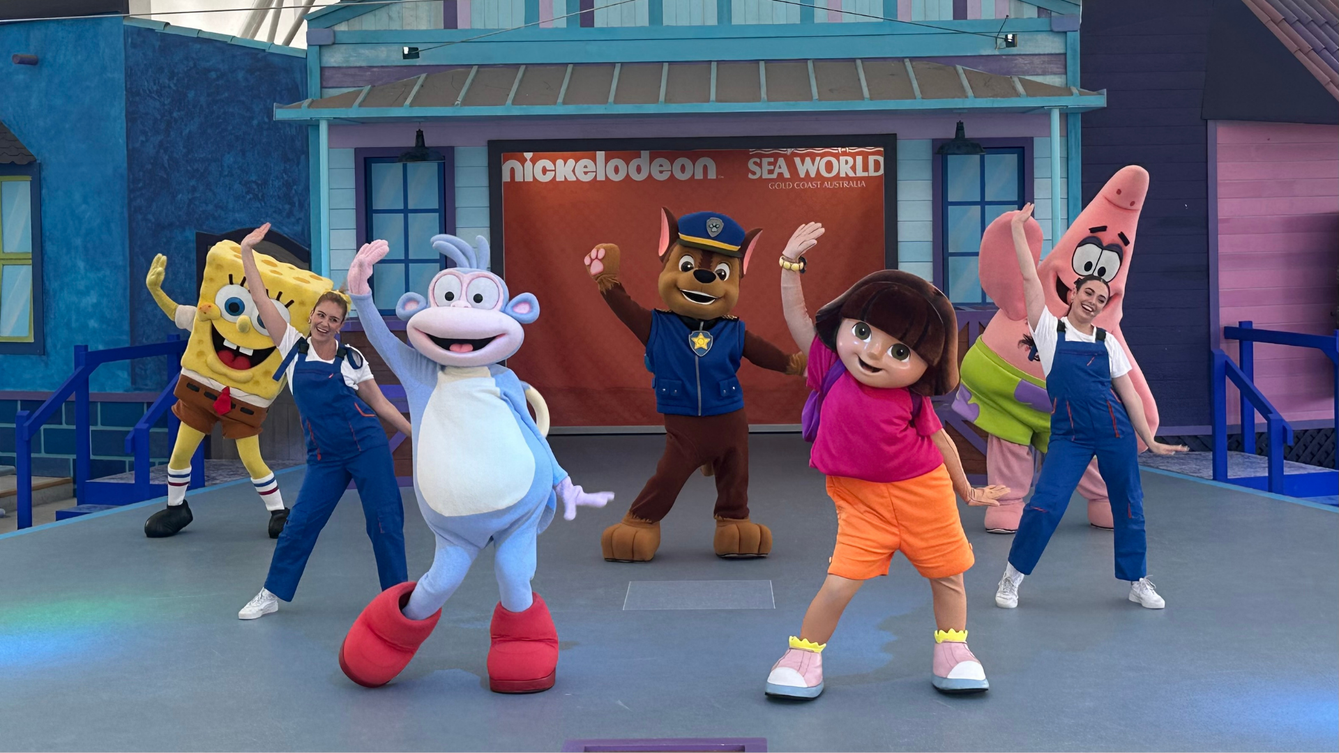 Costumed characters including SpongeBob, Boots, Chase, Dora, and Patrick dance on stage with performers in overalls at a Nickelodeon Sea World event, with colorful buildings in the background.