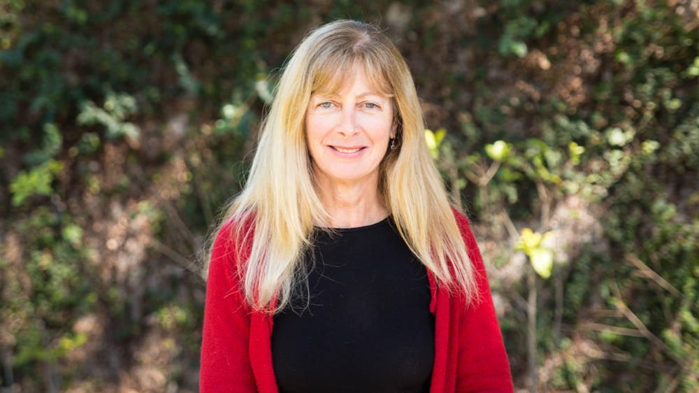 A woman with long blonde hair wearing a red cardigan and black top stands outdoors in front of leafy greenery.