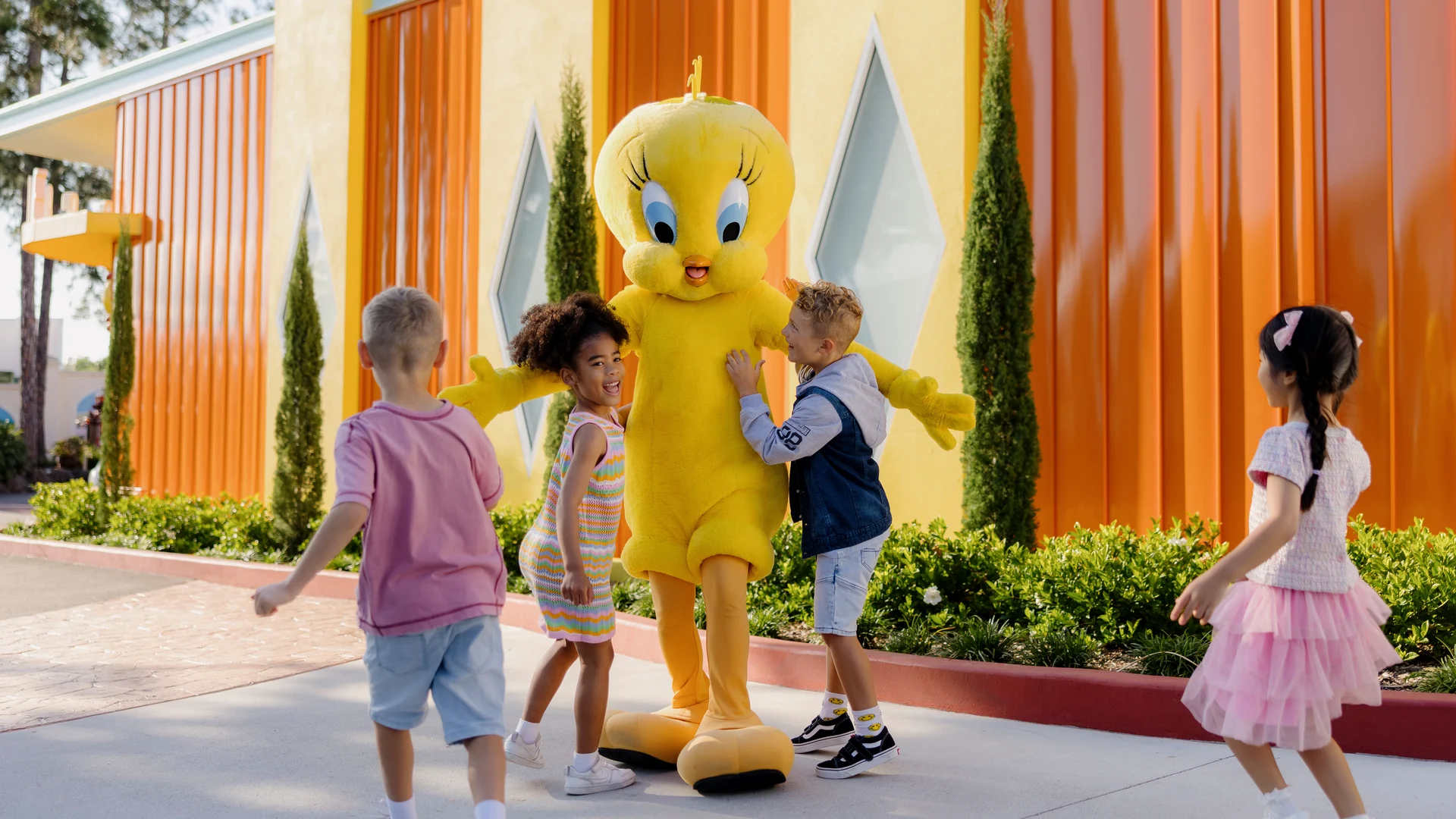 Four young children happily gather around and hug Tweety outside a colorful building with orange walls and geometric windows.