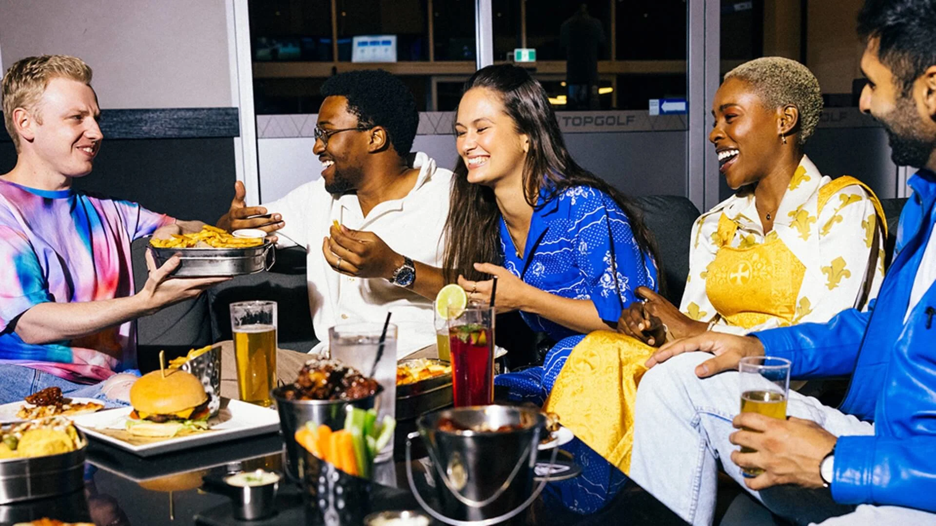 Group of friends laughing and sharing food and drinks at Topgolf Gold Coast, seated around a table with burgers, fries, and cocktails